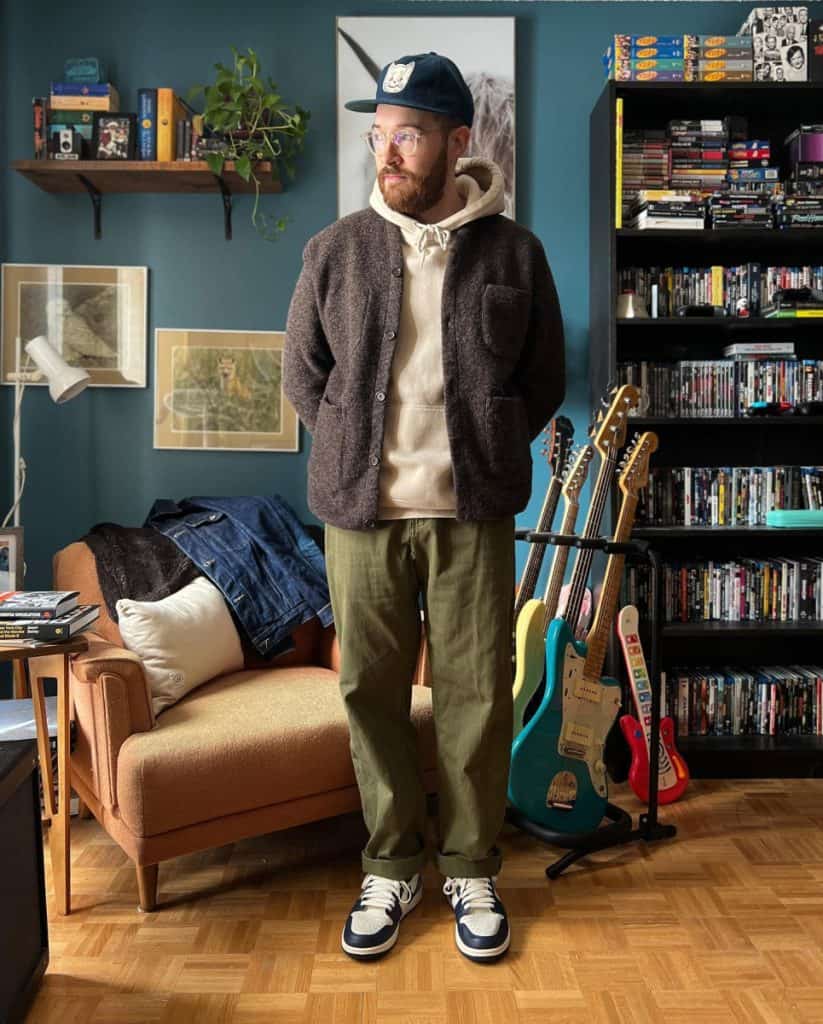 A man exudes casual sophistication in his home, wearing a brown cardigan over a hoodie with olive trousers, complemented by classic sneakers, a vintage cap, and a relaxed pose, embodying the perfect balance of comfort and style in men's fashion.