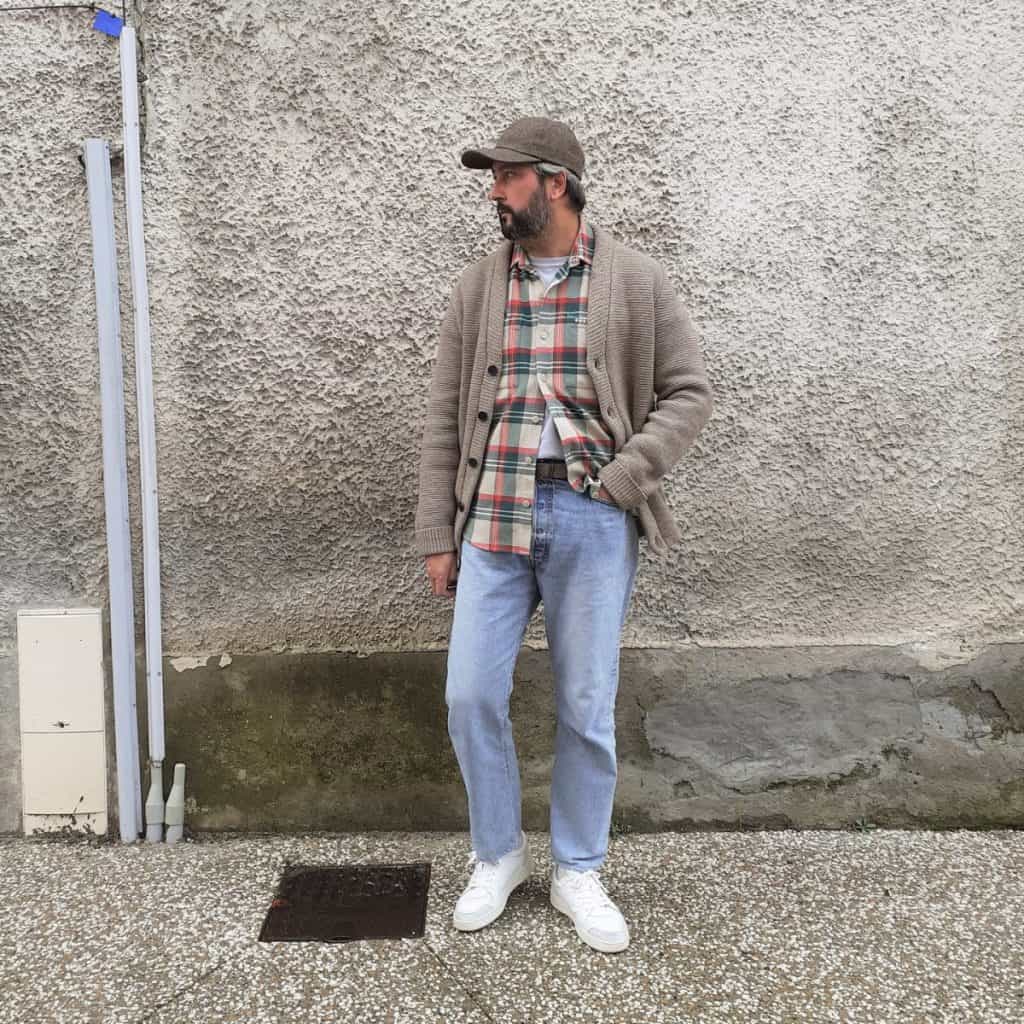 A man exudes effortless style in an urban setting, wearing a textured shawl collar cardigan over a plaid shirt, paired with light-wash jeans, a flat cap, and sleek white sneakers, perfectly blending traditional and modern elements in men's fashion.