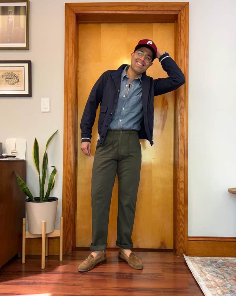A man leans in a doorway with a smile, sporting a navy button-up cardigan over a chambray shirt, paired with olive trousers and tan loafers, topped with a casual baseball cap, melding classic and contemporary elements in men's fashion.