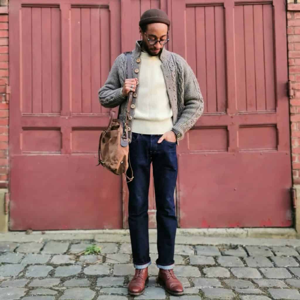 A stylish man poses thoughtfully outdoors, modeling a grey men's cardigan layered over a cream turtleneck, paired with dark blue jeans and brown leather boots, accessorized with a beret and a leather bag, exemplifying a refined approach to wearing a cardigan with jeans in men's fashion.