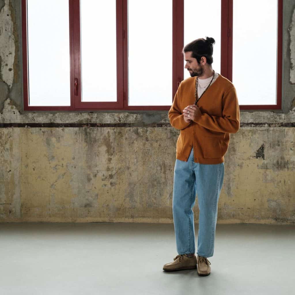 A man in a spacious room stands contemplatively, styled in a burnt orange men's cardigan sweater and light blue jeans, finished with desert boots, epitomizing a relaxed yet fashionable approach to wearing a cardigan sweater in men's fashion.