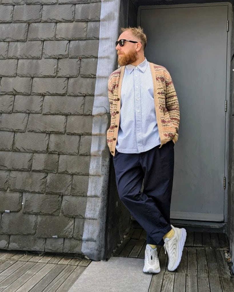 A stylish man leans against a grey wall, wearing a patterned cardigan over a light blue collar shirt with navy trousers and white sneakers, showcasing how to wear a collar shirt under a cardigan for a fashionable, casual men's look.