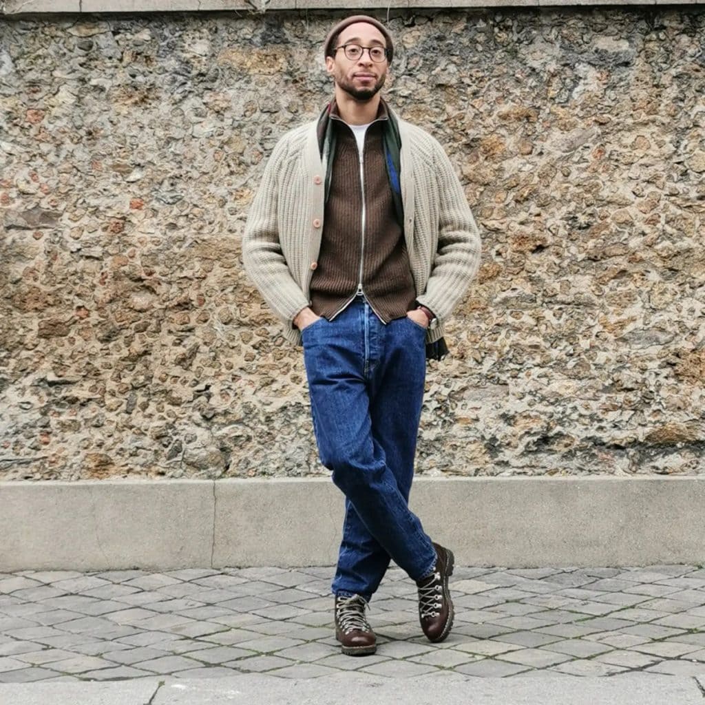 A man stands against a textured stone wall, wearing an oatmeal beige cardigan layered over a dark shirt, paired with blue jeans and brown boots, embodying a refined yet relaxed approach to men's cardigan fashion.