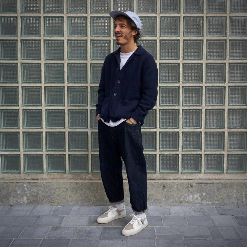 A man leans against a textured wall, styled in a classic navy cardigan over a white t-shirt, paired with dark jeans and white sneakers, reflecting a modern approach to wearing a navy cardigan in men's everyday fashion.
