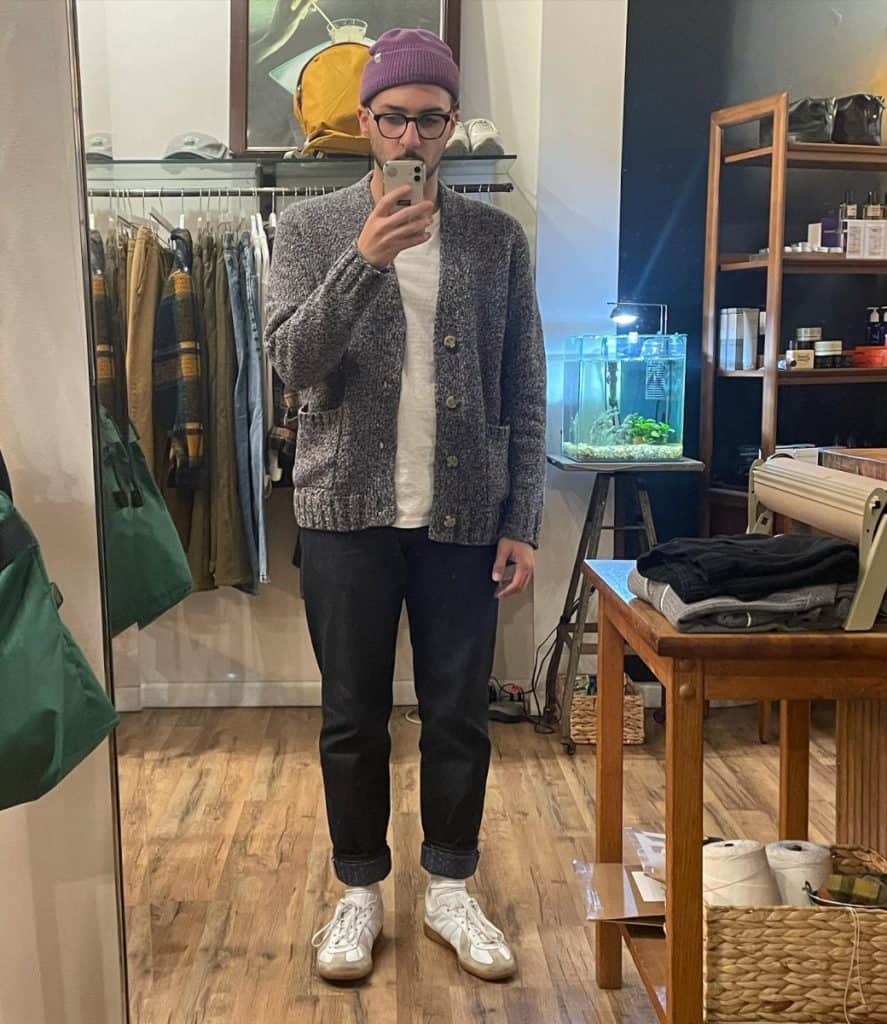 A stylish man in a boutique takes a selfie, wearing a textured grey cardigan paired with a white tee, cuffed jeans, white sneakers, and a purple beanie, showcasing a casual yet chic approach to men's cardigan fashion.