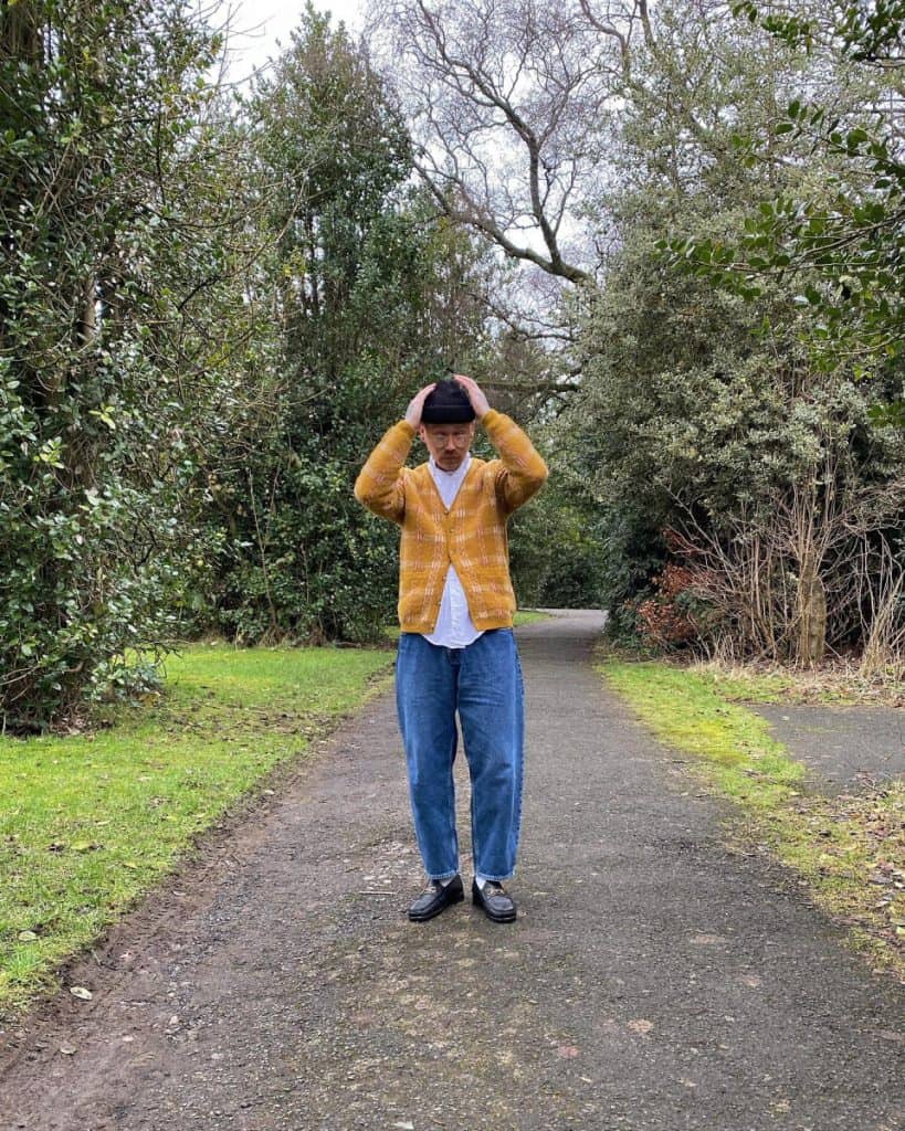A man stands on a woodland path, fashionably dressed in a bright yellow cardigan over a white shirt, paired with blue jeans and black loafers, showcasing how to stylishly incorporate a yellow cardigan into men's fashion.