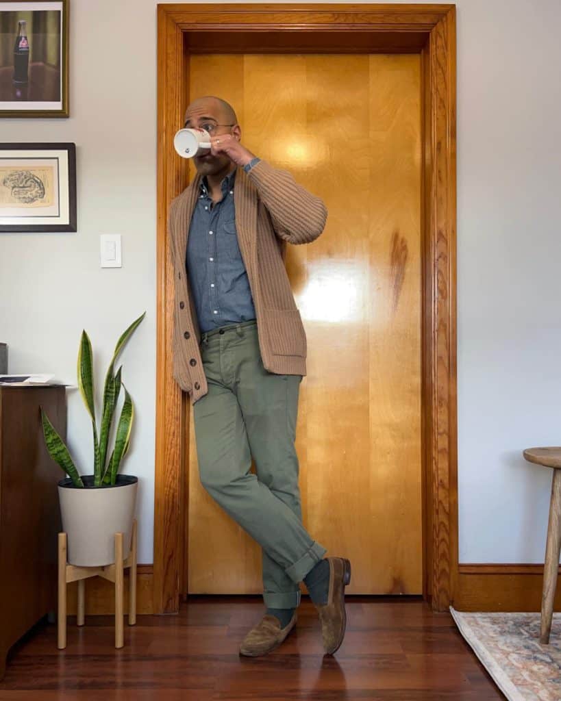 A stylish man takes a relaxed stance while sipping coffee, wearing a camel cardigan over a chambray shirt with rolled-up green trousers and suede boots, embodying a comfortable yet sophisticated approach to men's fashion with a cardigan.