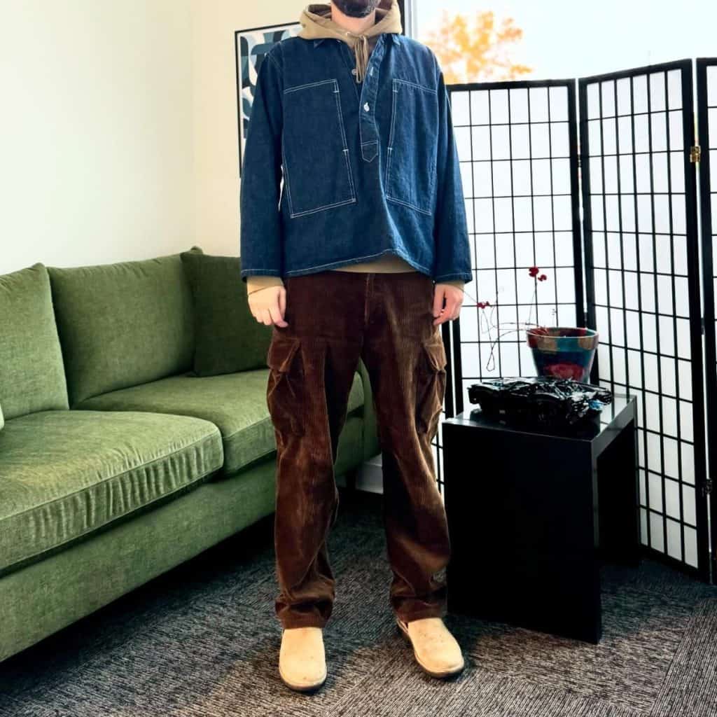 A man wearing a denim jacket over a hoodie paired with brown corduroy pants and suede boots, showcasing a textured, layered look.